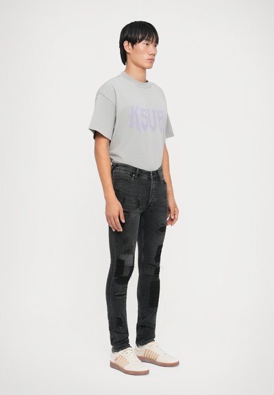 T-shirt grigia oversize con grafica viola, jeans skinny neri con strappi, e sneakers bianche con dettagli beige.