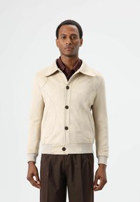 Beige suède jas met een gestructureerde kraag, knoopsluiting en decoratief stikwerk. Voorzien van zijzakken en geribde manchetten.