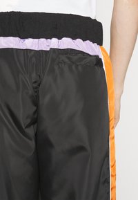 Černé syntetické tepláky s fialovým a oranžovým elastickým pasem a bočními akcenty, které mají malý detail zadní kapsy.