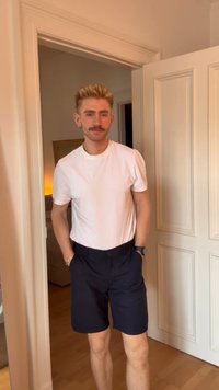 Young man with blonde hair and mustache standing in a doorway, wearing a white t-shirt, black shorts, and a wristwatch.