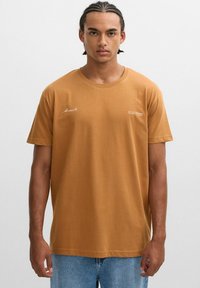 Camiseta de algodón marrón con mangas cortas y cuello redondo. Presenta texto impreso en blanco en el frontal. Combinada con jeans de mezclilla azul claro.