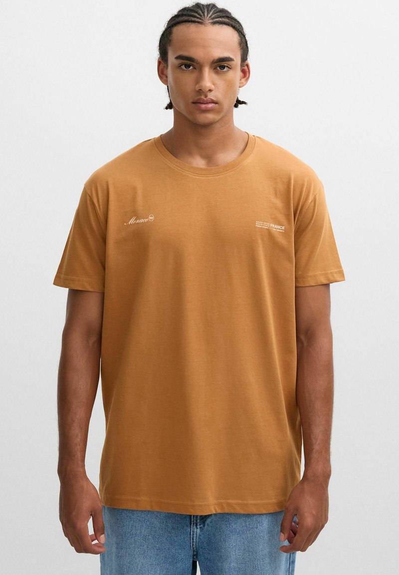 Camiseta de algodón marrón con mangas cortas y cuello redondo. Presenta texto impreso en blanco en el frontal. Combinada con jeans de mezclilla azul claro.