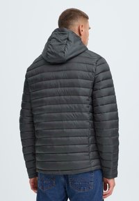 Grå, polstret jakke med vandret syninger, en aftagelig hætte og en glat tekstur. Sammen med blå denimjeans.
