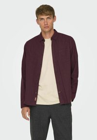 Bordeaux knappet skjorte i et tekstureret stof med en brystlomme, parret med en beige t-shirt og mørkegrå bukser.
