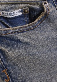 Τζιν τζιν με ανοιχτό μπλε ξεβαμμένο χρώμα, με κρυφή κλείσιμο με ορειχάλκινο κουμπί και ετικέτα με την ένδειξη "Juicy Couture Jeans" μέσα στη μέση της ζώνης.