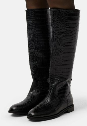 Botas negras hasta la rodilla con textura de cocodrilo, suelas planas, punteras ligeramente redondeadas, usadas por una persona de pie sobre una superficie blanca.