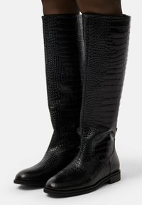 Schwarze knielange Stiefel mit Krokodilprägung, flachen Sohlen und leicht abgerundeten Zehen, getragen von einer Person, die auf einer weißen Oberfläche steht.