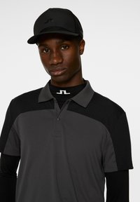 Grå och svart pikétröja med krage och korta ärmar, med en knapplista fram. Matchad med en svart keps som visar en logotyp.