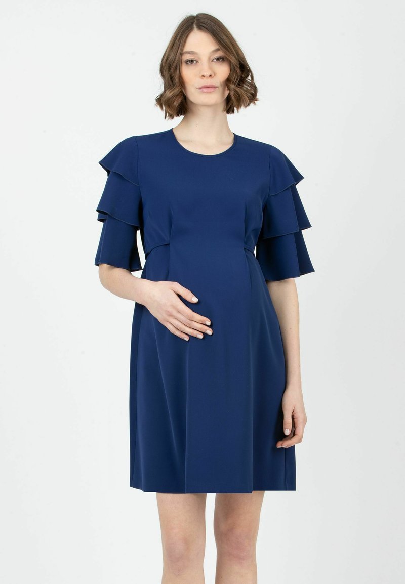 Äitiysmekko tummansinisestä, sileästä kankaasta. Ominaisuuksiin kuuluu kerrokselliset volanttisangat ja  vartalonmyötäinen yläosa, joka on suunniteltu tukemaan kasvavaa vatsaa.