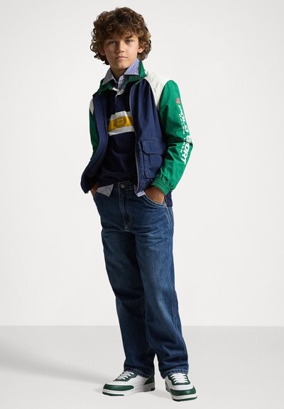 Casaco azul-marinho e verde, camisa às riscas e jeans azuis. Ténis brancos e verdes. Look casual com acentos e texturas desportivas distintas.