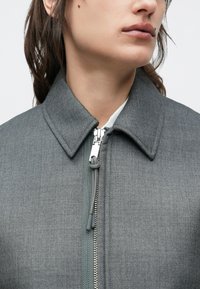 Žena v sivej bunde so zipsom a veľkým golierom, ktorá ukazuje časť tváre a krku na jednoliatom pozadí.