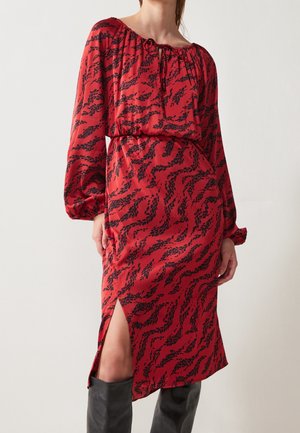 Femme portant une robe midi rouge à manches longues avec des motifs de feuilles noirs, une fente latérale et des bottes noires hautes jusqu'aux genoux.