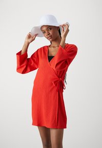 Vestido rojo ajustado con tejido texturizado, escote en forma de v y detalle de lazo en la cintura. Modelo con un sombrero blanco de ala ancha, posando frente a un fondo claro.