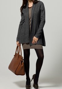 Cardigan grigio a coste con apertura frontale, abbinato a un vestito beige, collant neri e borsa in pelle marrone. Le scarpe col tacco completano il look.