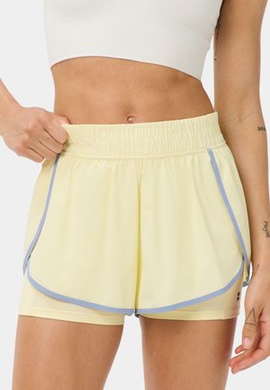 Vrouw draagt lichtgele sportshorts met lichtblauwe rand en een wit kort topje, houdt de tailleband van de shorts vast.