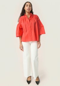 Blusa rossa di cotone con maniche a palloncino e scollatura arricciata, abbinata a pantaloni bianchi a gamba larga e décolleté neri a punta.