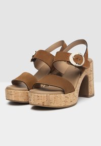 Sandales en suède marron avec deux larges brides et une grande boucle ronde, dotées d'une plateforme en liège et d'un talon carré. Doublure intérieure lisse.