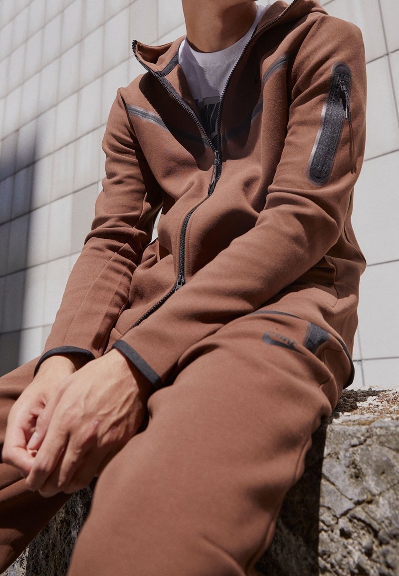 Braune Jacke mit Reißverschluss und Kapuze sowie einer Reißverschlusstasche, die kontrastierende schwarze Akzente und einen weichen, strukturierten Stoff aufweist. Getragen mit passenden Hosen.