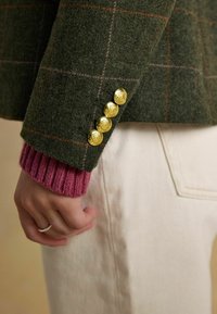 Vihreä villatakki, jossa on ruutukuosi, kullanväriset napit hihoissa ja vaaleanpunainen neuleristetus. Yhdistetty valkoisiin kangashousuihin.