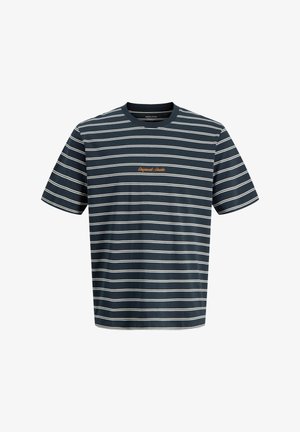 Smilende dreng stående iført en mørkeblå og hvidstribet t-shirt, sorte bukser og hvide sneakers på en ensfarvet lys baggrund.