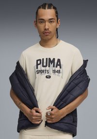 Puma dunvest i mørkeblå med quiltet struktur, båret åben over en cremefarvet T-shirt med fedt "PUMA SPORTS 1948" print.
