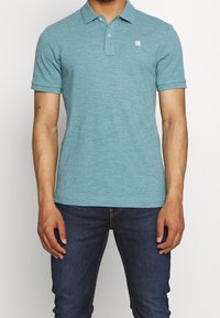 Polo couleur sarcelle, fabriqué en tissu léger. Il présente un col à revers, des manches courtes et un petit logo contrastant sur la poitrine.