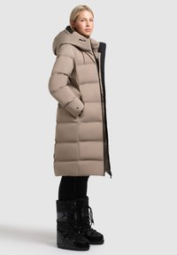 Beige gewatteerde lange jas met een grote capuchon, rits aan de voorkant en zijzakken, gedragen over zwarte leggings en zwarte winterlaarzen.