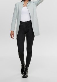 Abrigo azul claro con un acabado texturizado, llevado sobre una blusa blanca y combinado con jeans skinny negros y botas negras de cordones.