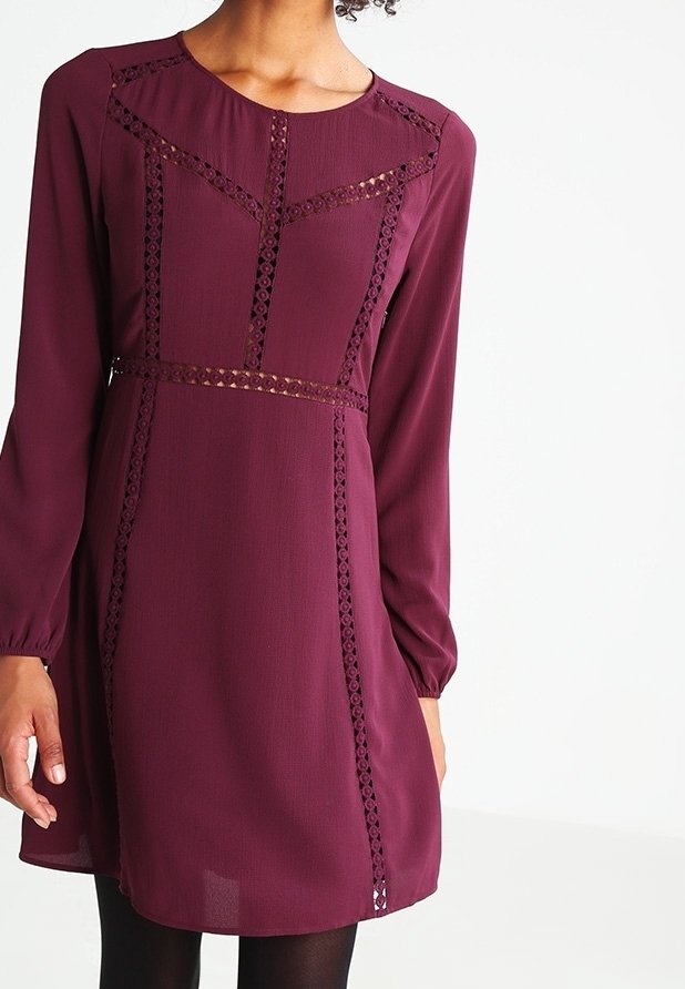 Femme portant une robe bordeaux à manches longues avec des détails en dentelle sur la poitrine, la taille et les coutures verticales, associée à des collants noirs.