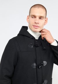 Abrigo tipo duffle negro con capucha, que presenta cierres de toggle y una textura similar a la lana. El modelo lleva una camiseta de cuello alto blanca debajo.