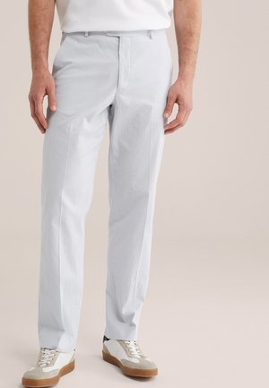 Homme portant un pantalon gris clair texturé, des baskets blanches avec des accents beige et une chemise blanche rentrée, sur un fond uni.