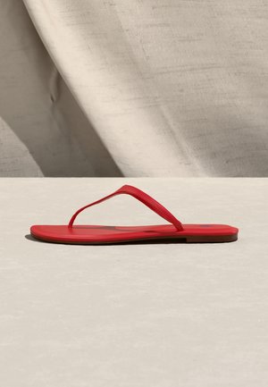 Flat red flip-flop sandal with thin straps placed on a light beige surface against a textured beige fabric background.