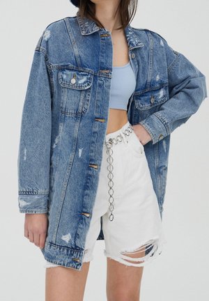 Jeune femme portant une veste en jean bleue délavée oversize, un crop top bleu clair, un short blanc déchiré et une ceinture chaîne argentée à gros maillons.