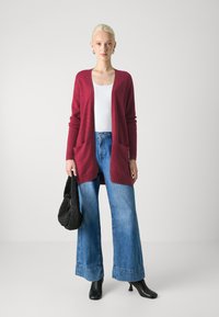 Burgundy lång cardigan med fickor, bärs över en vit topp, tillsammans med ljusblå vida jeans och svarta klackboots. Svart vävd väska.