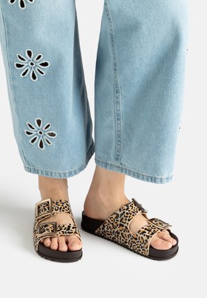 Feet wearing leopard print slide sandals with double buckle straps, paired with light blue wide-leg jeans featuring flower cutout designs.