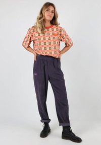 T-shirt à motif orange avec des manches courtes ; pantalon en velours côtelé bleu marine avec une marque violette ; personne portant des bottes noires à lacets.