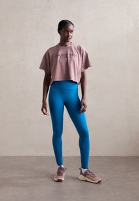 Rövidített mályva színű póló, rajta a "PROJECT SOCK" felirattal, magas derekú türkiz leggings, és bézs színű, textúrázott felületű sneaker cipők.
