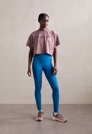 Skraćeni lila t-shirt s natpisom "PROJECT SOCK", visoko strukirane tirkizne tajice i bež tenisice s teksturiranom površinom.