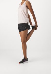 Camiseta deportiva de color rosa claro, pantalones cortos negros con logo y zapatillas de correr negras con acentos en naranja y púrpura. Individuo estirando una pierna.