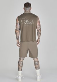 Ärmelloses olivgrünes Tanktop mit weißem Logo auf dem Rücken, kombiniert mit passenden braunen Shorts. Verziert mit Tattoos an den Armen und modernen weißen Schuhen.