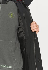 Chaqueta exterior negra con cierre en la parte delantera, que presenta un sudadera gris debajo con un logo y bolsillo. El interior tiene un forro negro suave.