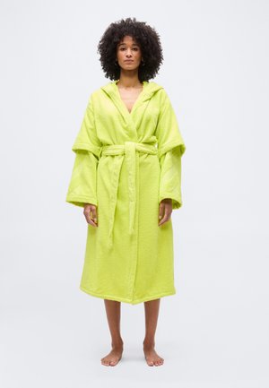 Woman with curly hair stands barefoot wearing a lime green hooded bathrobe with wide sleeves and a tied belt against a plain white background.