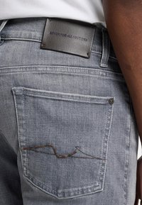 Jeans in denim grigio con una tasca sul retro con un caratteristico motivo di cucitura marrone e un'etichetta in pelle nera del brand in vita.