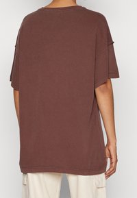 Camisa de algodón marrón de gran tamaño, con un cuello redondo, hombros caídos y mangas cortas con bordes sin rematar.
