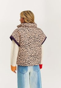 Krótka kamizelka puchowa z krótkim rękawem w jasnoróżowym kolorze z czarnymi cętkami w panterkę, z wysokim kołnierzem i fioletowymi wykończeniami. Nałożona na kremowy sweter z kolorowymi mankietami.