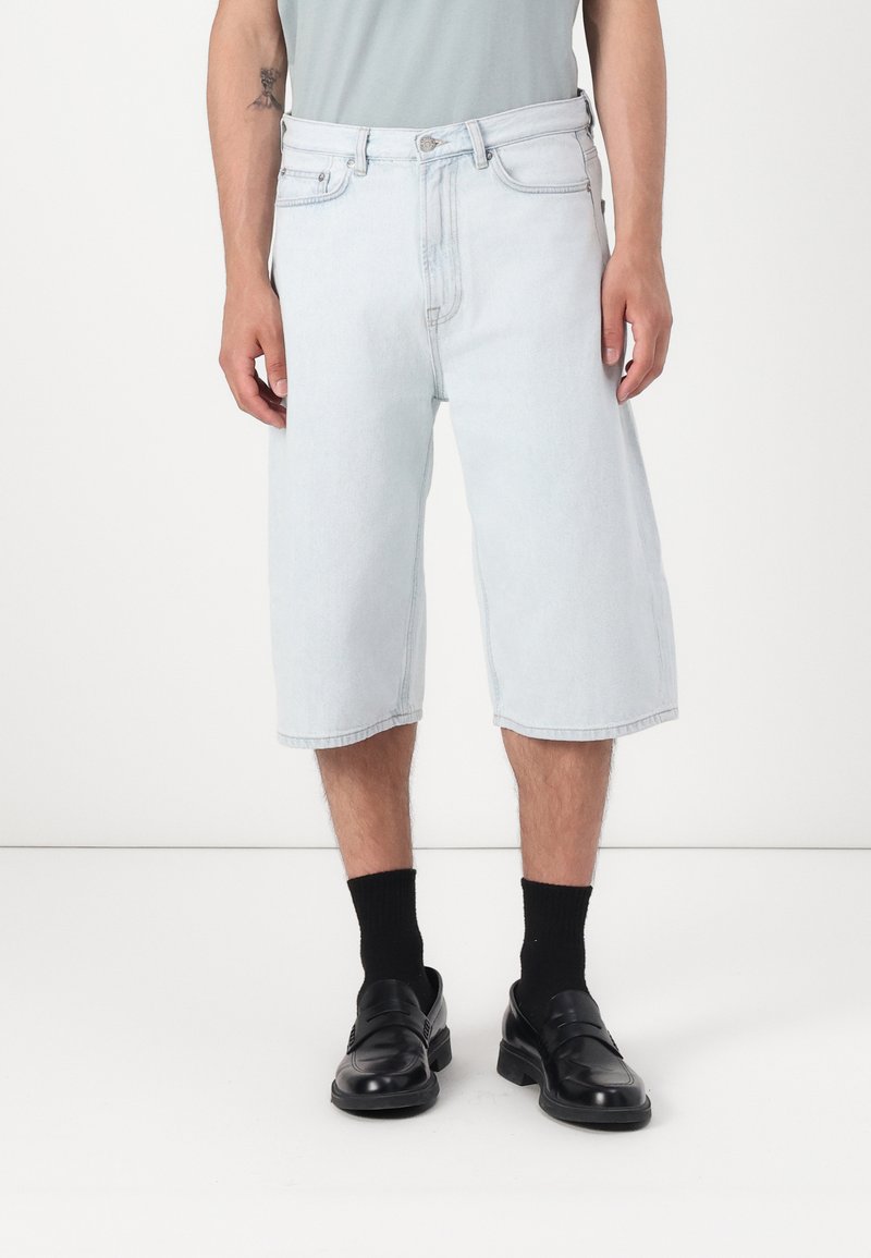 Shorts de mezclilla azul claro con un corte relajado, alcanzando la mitad de la pantorrilla, con cinco bolsillos y cierre de botón. Combinados con zapatos y calcetines negros.