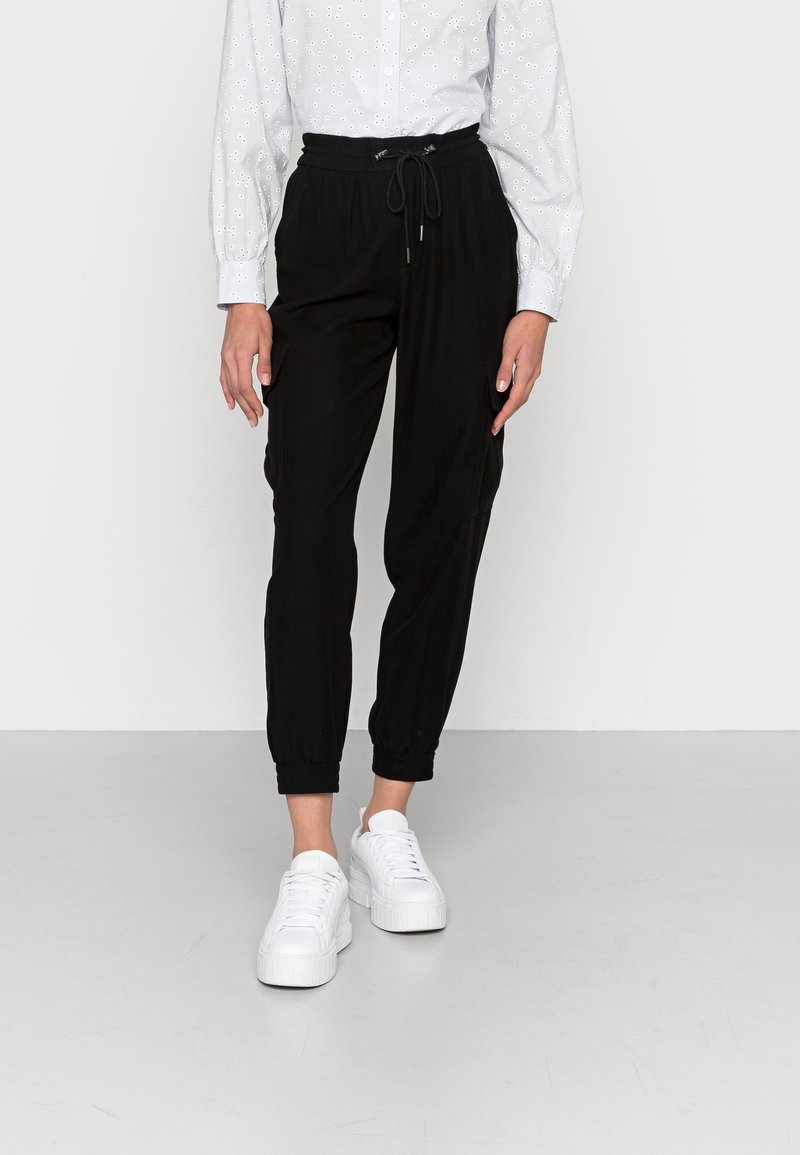 Personne portant un pantalon de jogging noir à cordon, une chemise blanche à motifs à manches longues et des baskets compensées blanches, debout sur un sol gris.