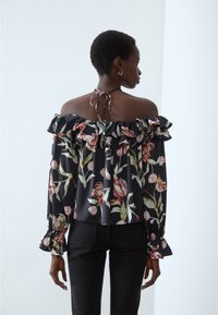Black floral off-shoulder blouse with ruffles, featuring a tie-back design, long sleeves, and a loose fit adorned with multicolored floral patterns.