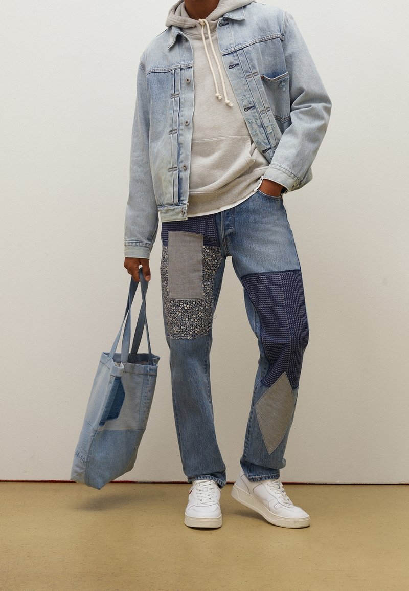 Denim jacket and patchwork jeans in various shades of blue, featuring mixed textures and a floral pattern. Light gray hoodie and tote bag.