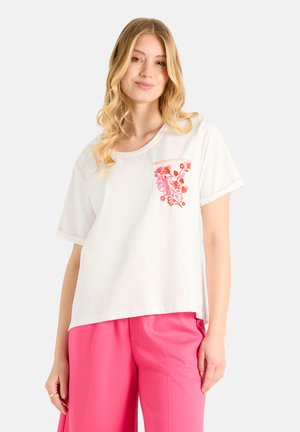 Junge Frau mit langen blonden Haaren, die ein weißes kurzärmeliges Oberteil mit roter Blumenstickerei und knallpinke Hose trägt, steht vor weißem Hintergrund.